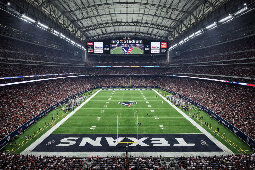 Wide-angle view of NRG Stadium in Houston, Texas, showing the Houston Texans football field under the closed retractable roof, with a packed crowd, bright green turf, end zone branding, and the stadium’s massive video scoreboard during an NFL game.