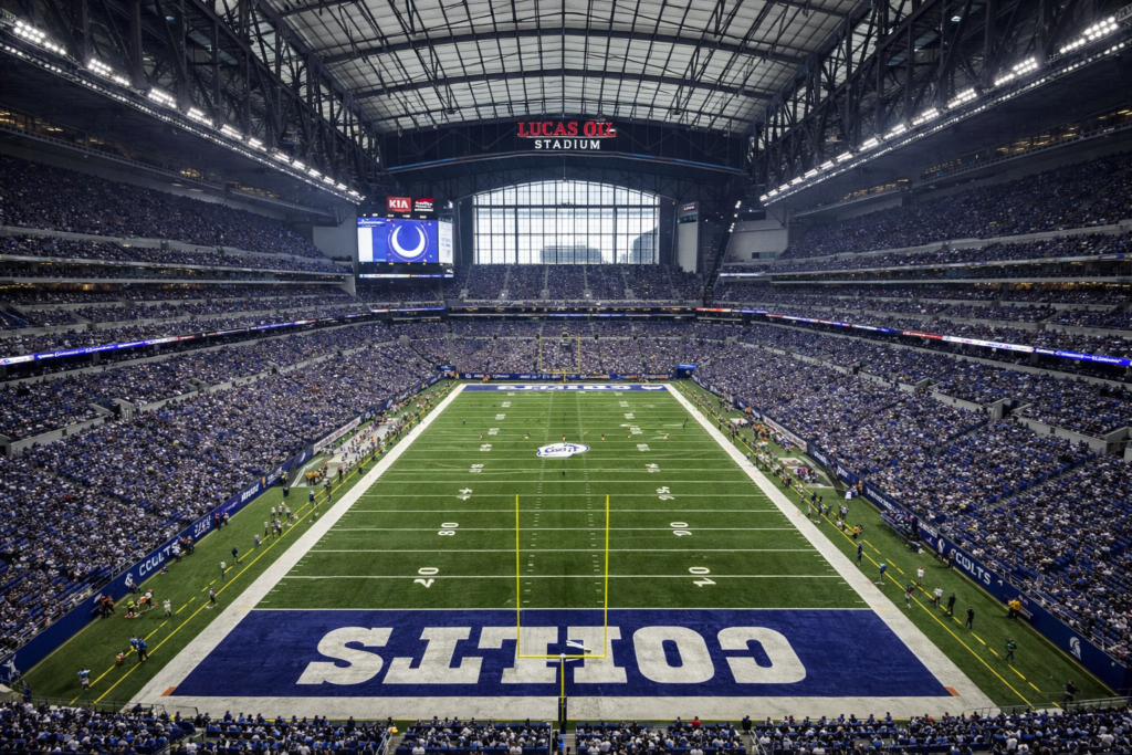 Wide-angle interior view of Lucas Oil Stadium in Indianapolis during an NFL game, showing the full football field, Colts end zones, retractable roof structure, packed stands, and stadium architecture.